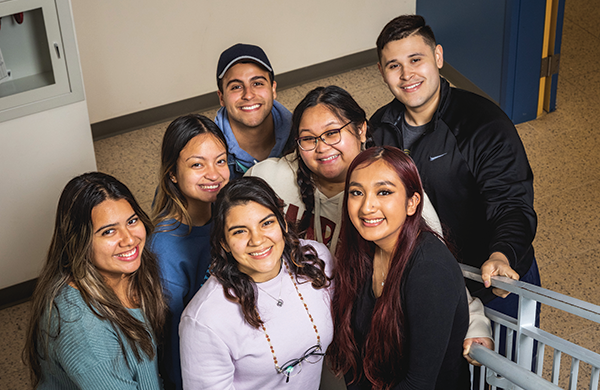 Seven students standing at the bottom of a staircase, smiling up at the camera.