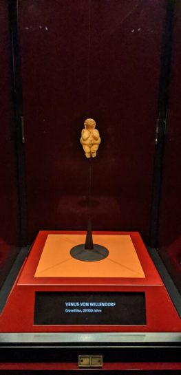 The Venus of Willendorf sitting in her glass case on a black background. The museum label reads "Venus von Willendorf."